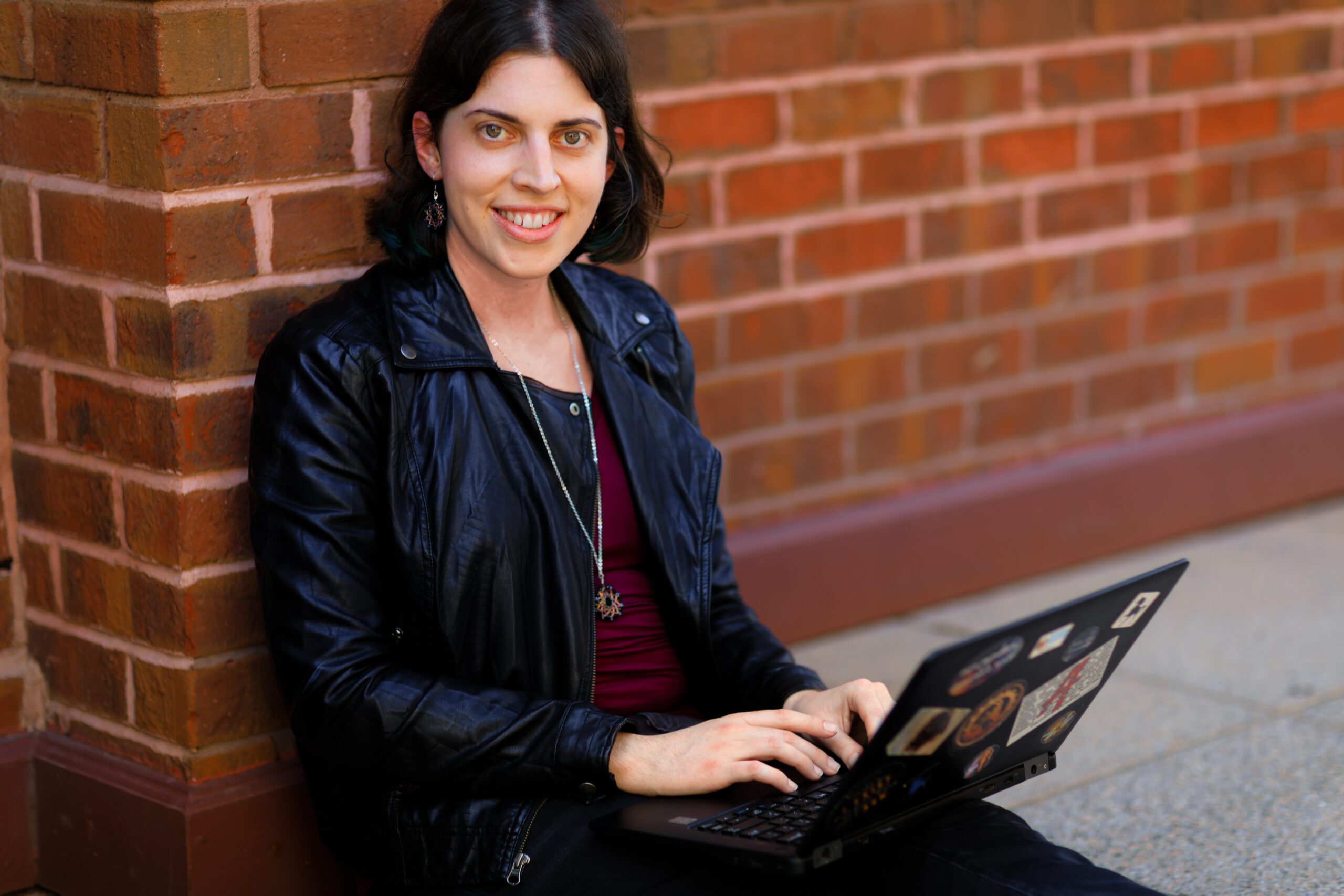 The image is of author coach and book business strategist Janeen Ippolito sitting on the ground and leading against a brick wall with her laptop in her lap. She's smiling at the viewer.